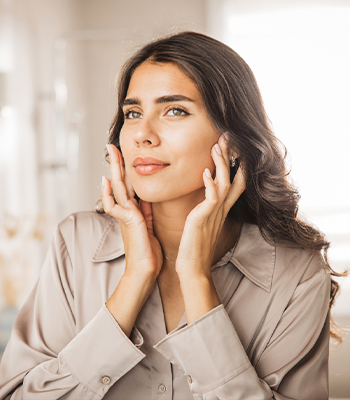 Skincare portrait of a woman touching her face, long dark hair, neutral blouse, soft natural light