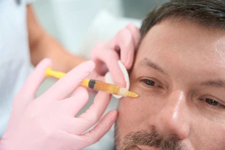 Close-up of a man receiving cosmetic tear-trough filler injection near the eye from a clinician in pink gloves