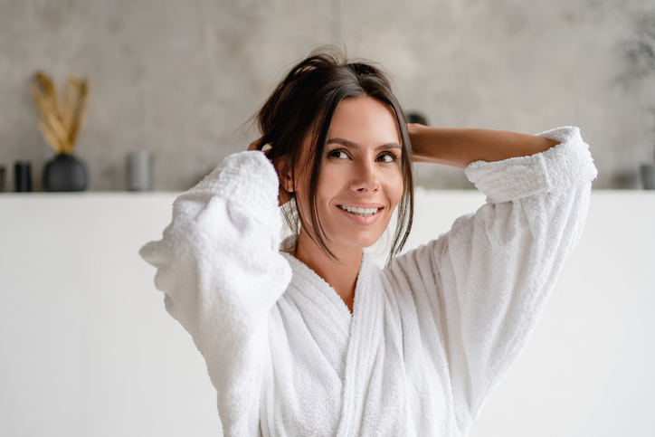 A smiling woman in a white bathrobe adjusting her hair in a bright, modern bathroom.