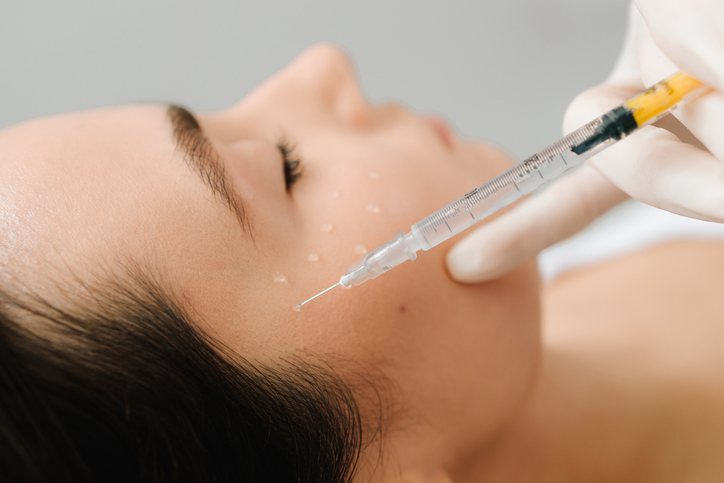 A woman receiving a cosmetic facial injection in the cheek from a gloved clinician