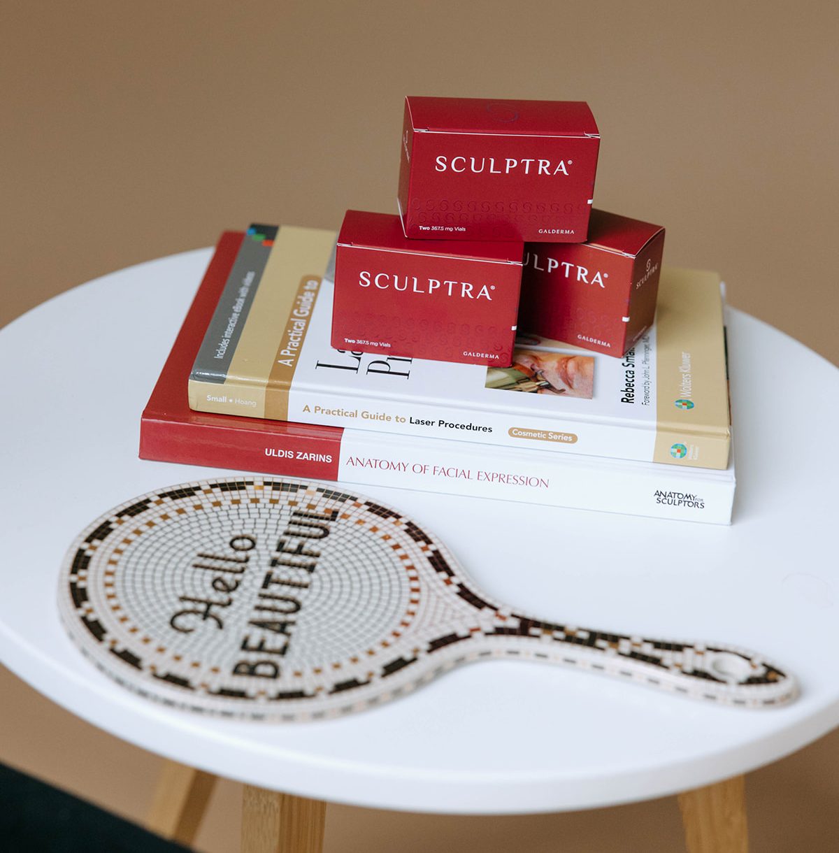 Sculptra product boxes stacked on aesthetic books beside a mosaic hand mirror reading 'Hello Beautiful' on white table