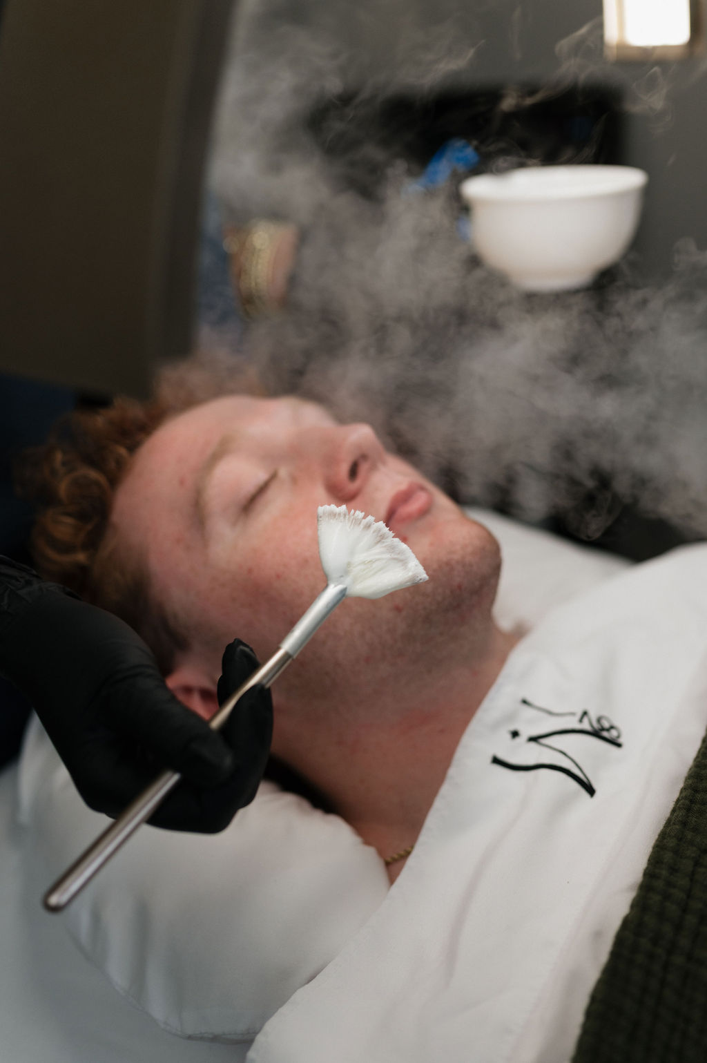 A man receiving a spa facial with a fan brush amid facial steam as an esthetician applies skincare