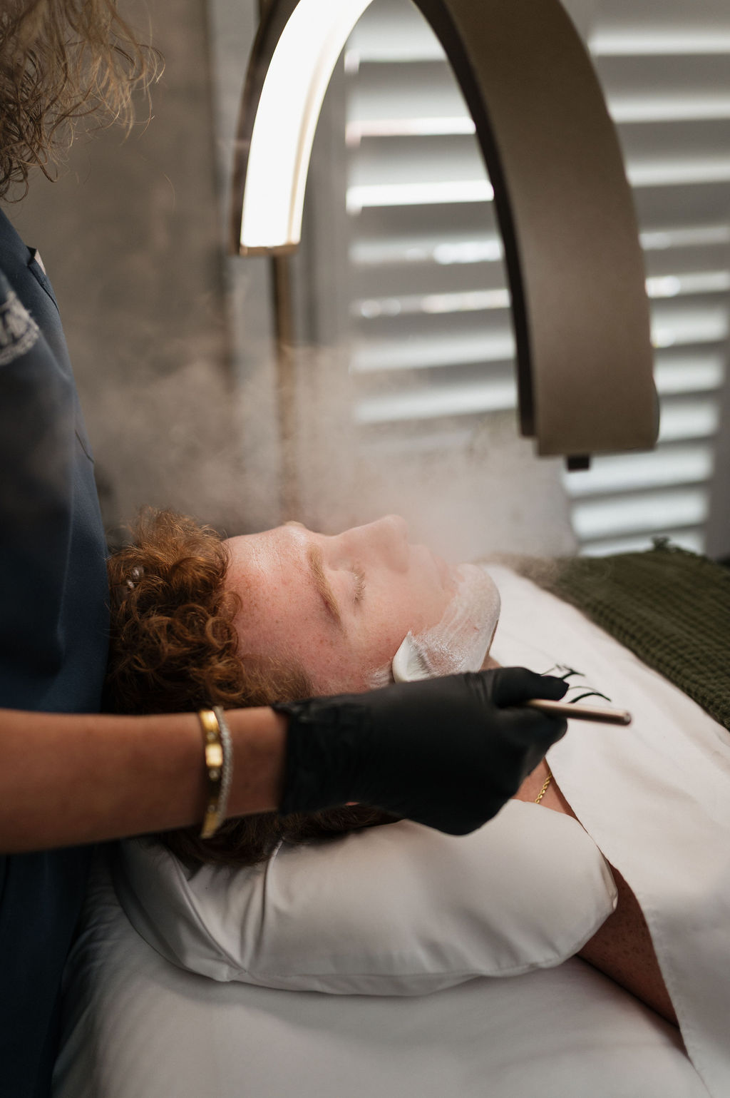 A man receiving a steam facial while an esthetician applies a skincare mask at a spa