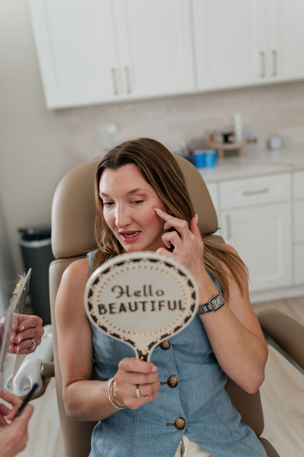 A woman in a cosmetic clinic chair examining her cheek with a hand mirror reading 'Hello BEAUTIFUL'