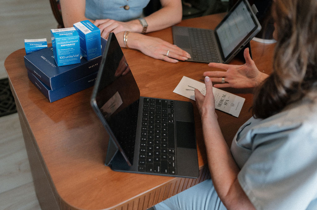 A woman consults with a provider at a medical spa, laptops open and boxes of injectable product on the table