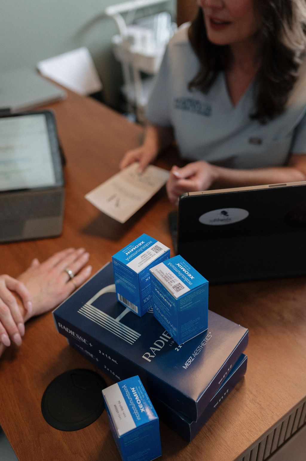 Woman in scrubs consulting a client over Radiesse dermal filler and blue injectable boxes on a wooden table.