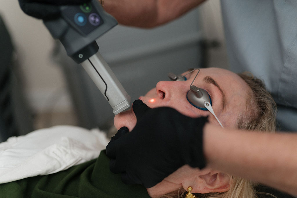 Woman receiving laser facial treatment as tech in black gloves holds laser at her cheek; she wears eye shields.