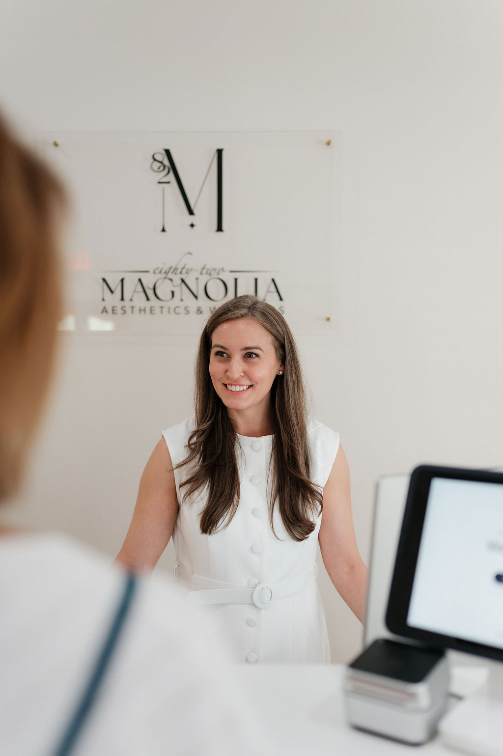 Smiling woman receptionist at Magnolia Aesthetics front desk in white dress with computer