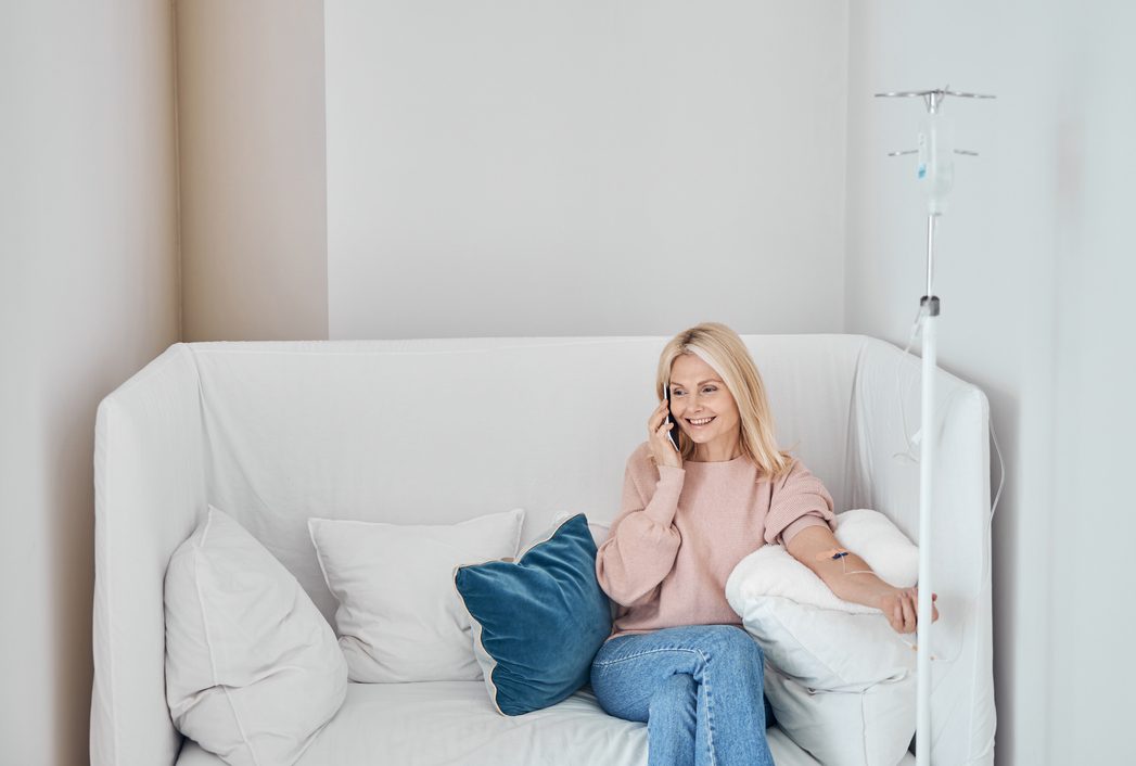 A woman receiving an IV infusion at home on a white couch, smiling while talking on her phone.