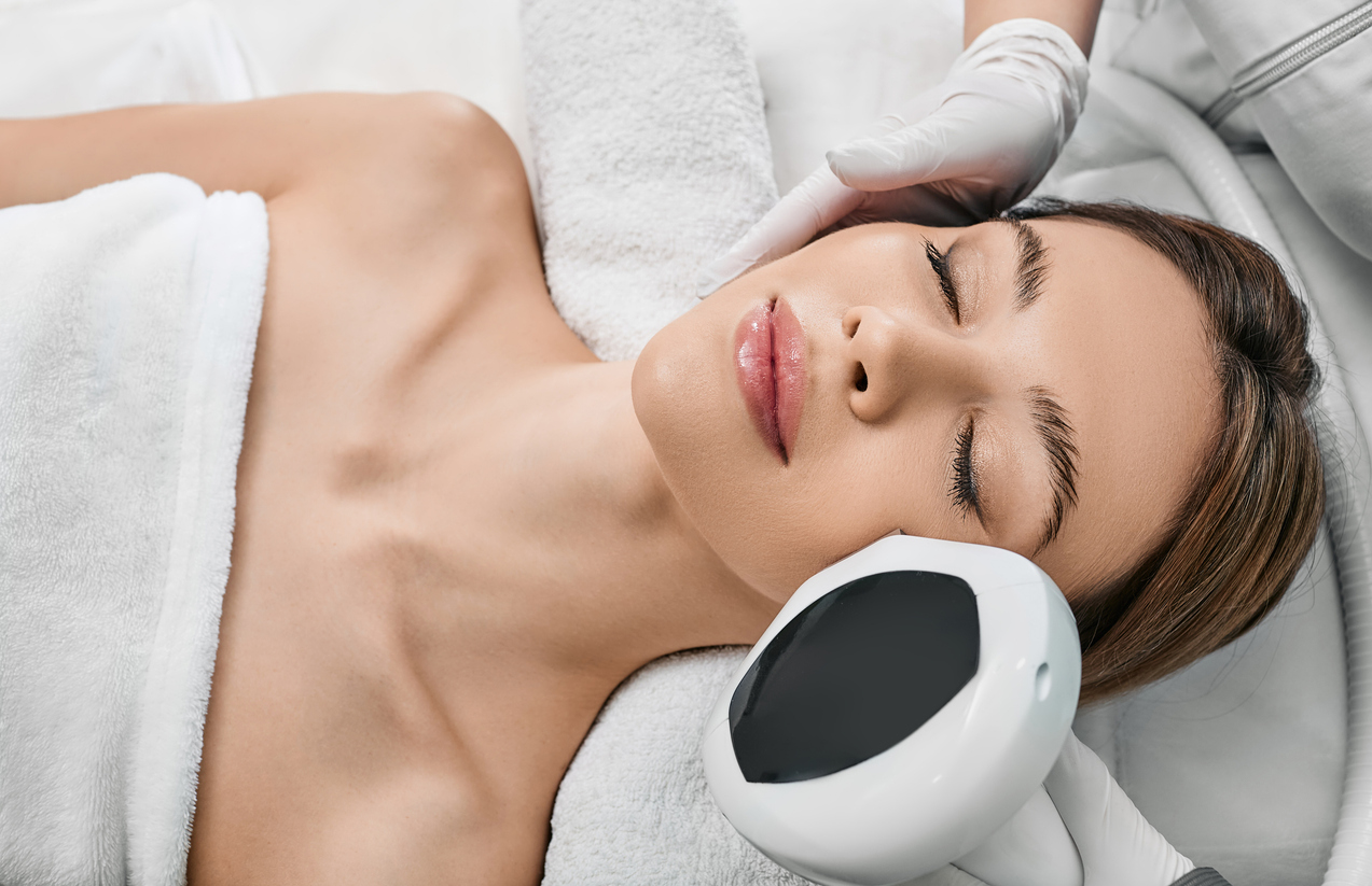 A woman receiving a professional facial laser treatment with handheld device at a spa, eyes closed, relaxed.