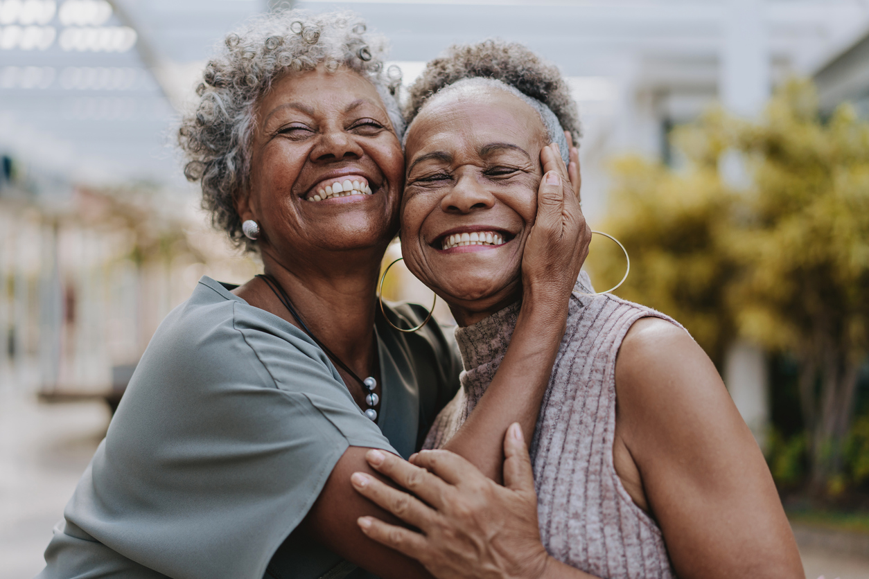 Two older women smiling and hugging closely outdoors, joyful friends sharing a warm embrace.