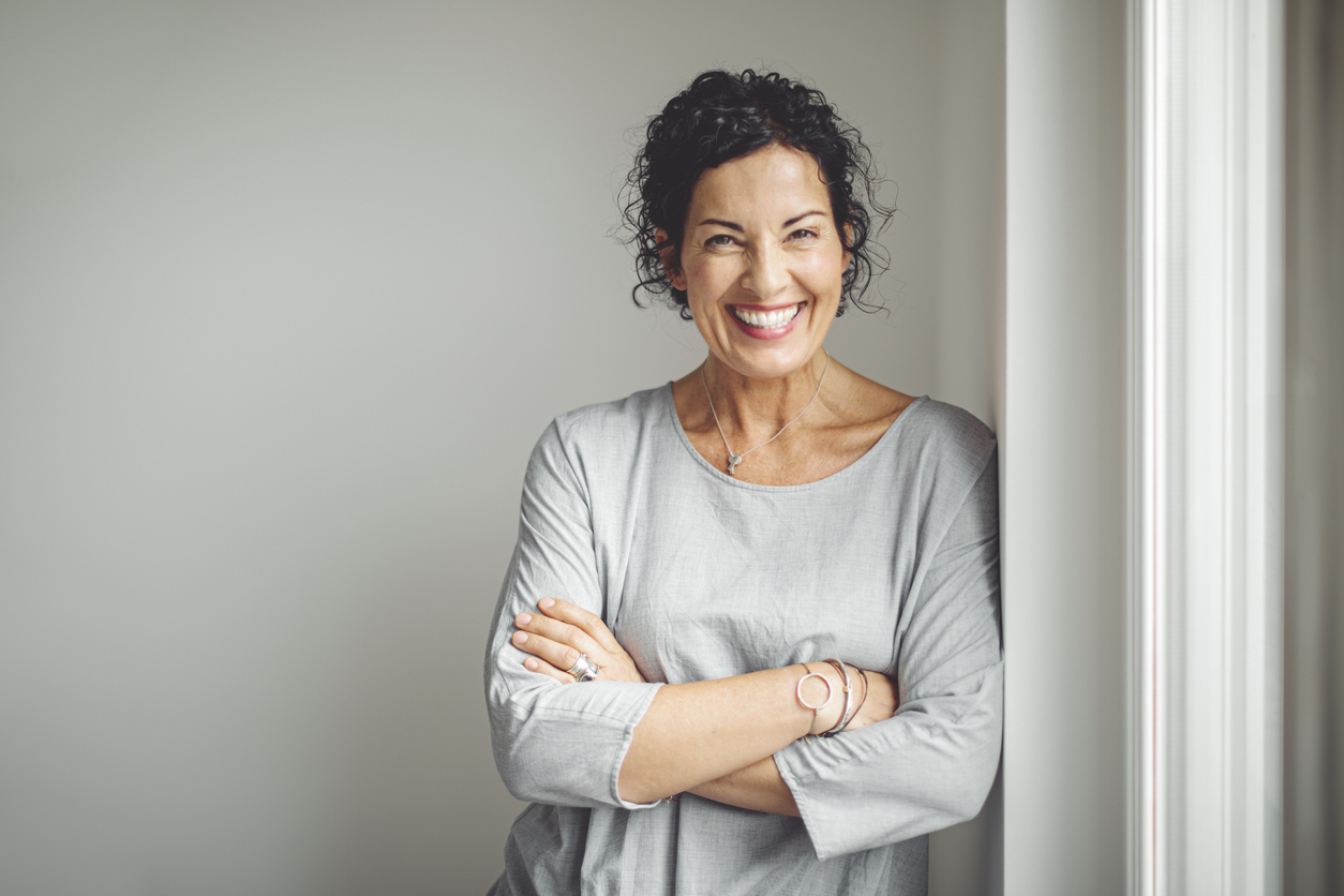 Smiling woman in gray blouse leaning against wall with arms crossed, confident portrait