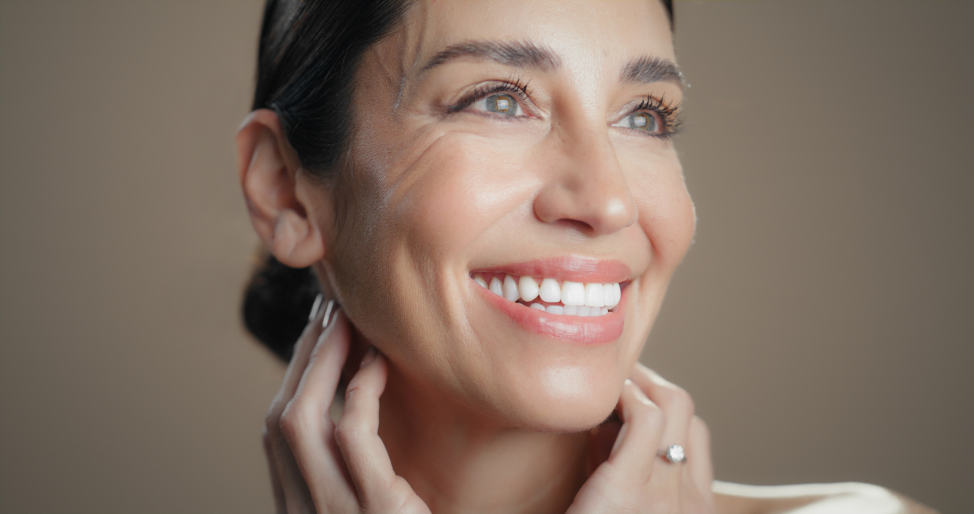 Close-up of a smiling woman with radiant skin, bright white teeth, and hands near her neck wearing a ring.