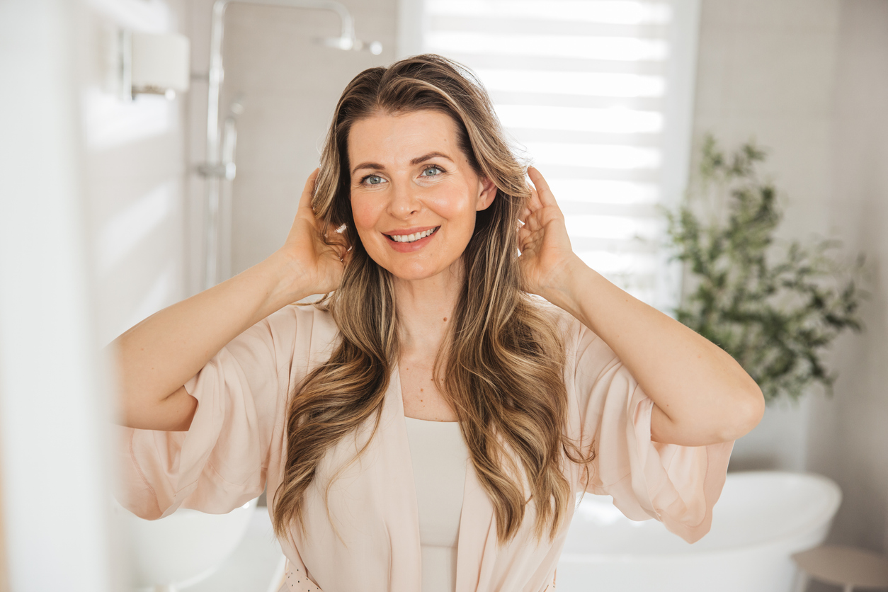 Smiling woman with long wavy hair adjusting hair in bright bathroom, natural skincare morning routine