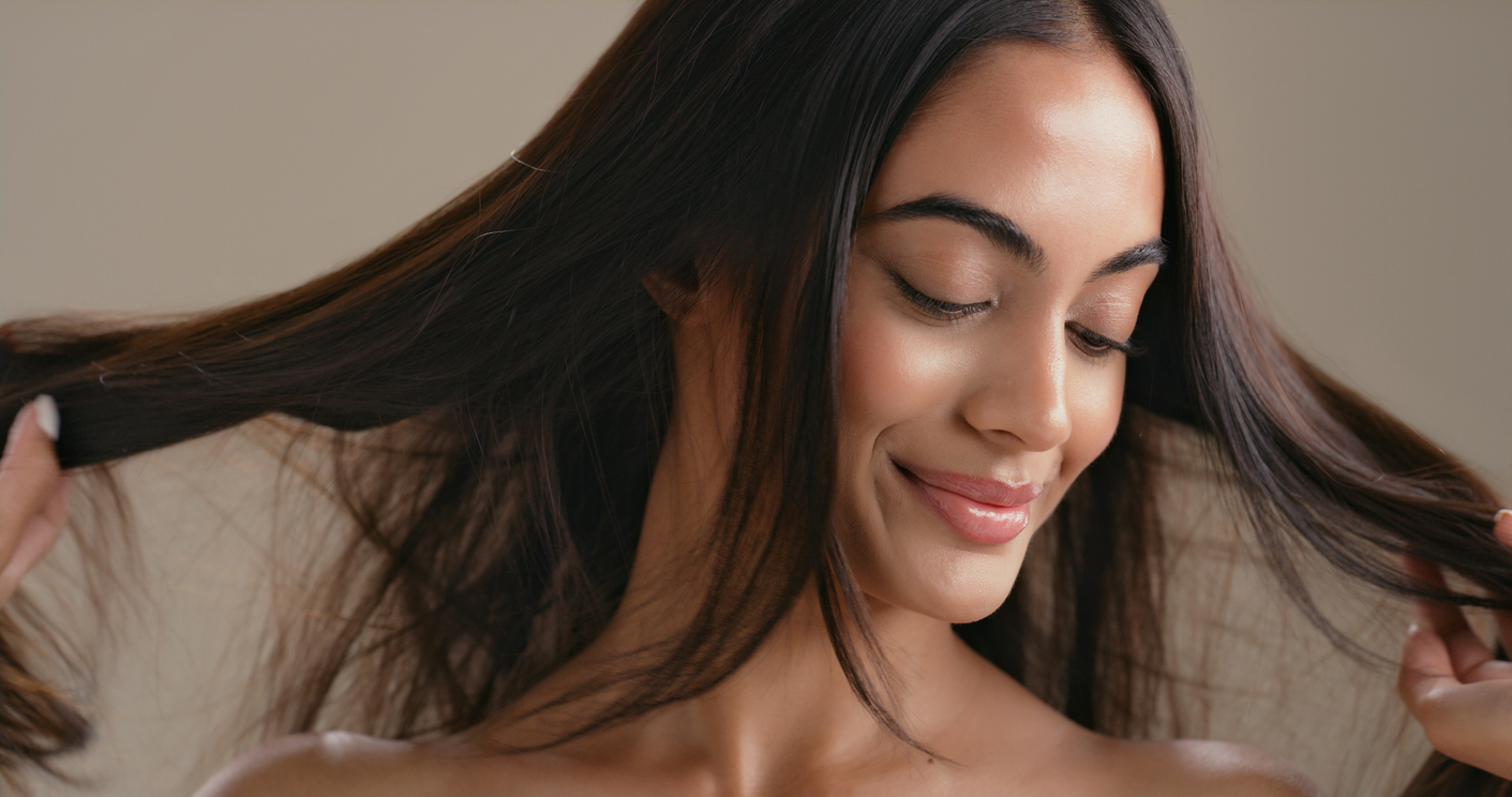 A woman with long dark hair smiling gently while holding her hair, showcasing smooth glowing skin