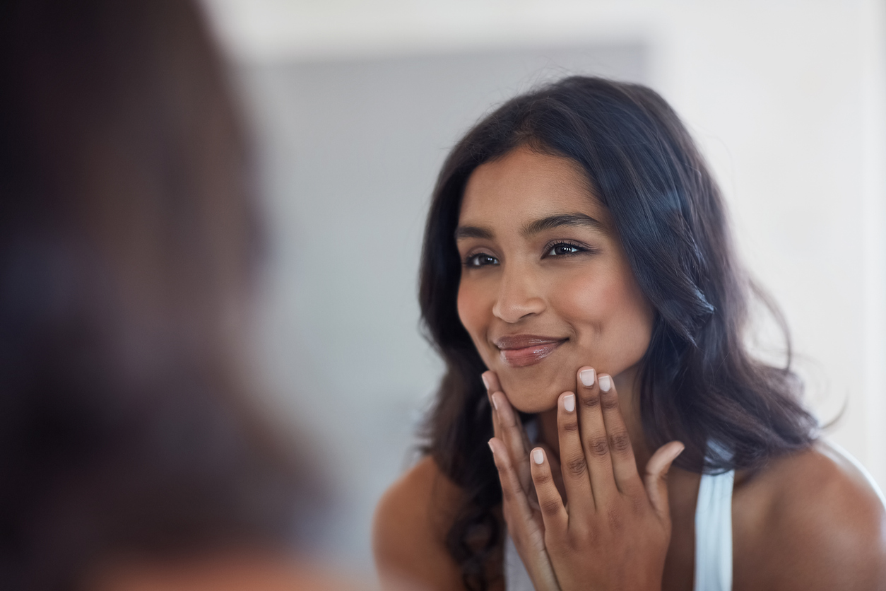 A woman smiling while touching her face in front of a mirror, showcasing glowing clear skin