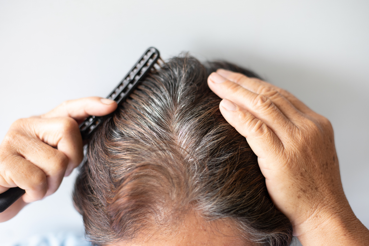 A person combing gray-streaked hair at the scalp, showing a center part and slight thinning.